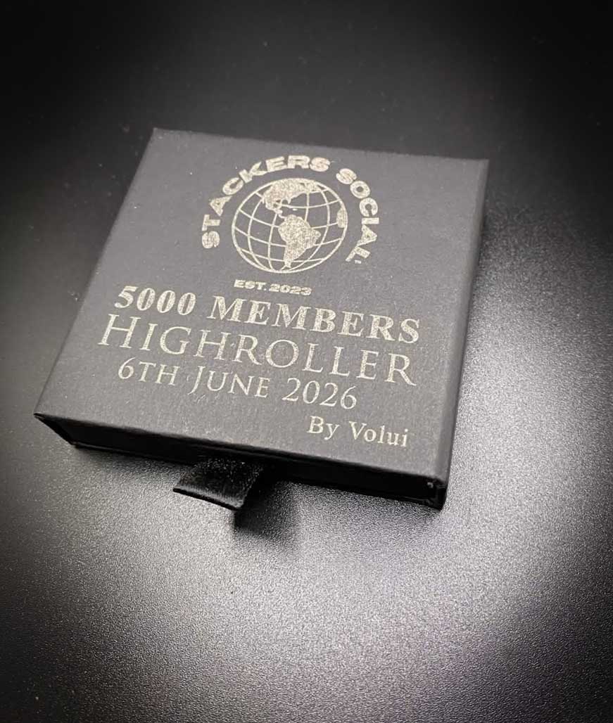 A square black presentation box with a pull tab, featuring white printed text and a globe logo that reads 'Stackers Social' and '5000 Members Highroller 6th June 2026 By Volui', photographed on a dark textured surface.