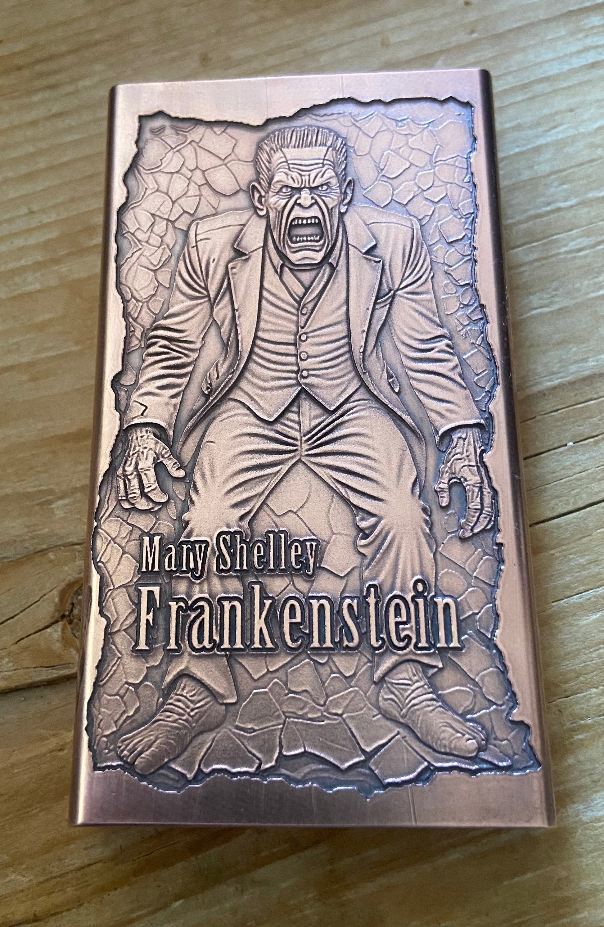 A rectangular metal box featuring an embossed illustration of Mary Shelley as Frankenstein, placed on a wooden surface. The box has a metallic rose-gold or copper-toned finish with gray engraved artwork and the text 'Mary Shelley Frankenstein' on the front.