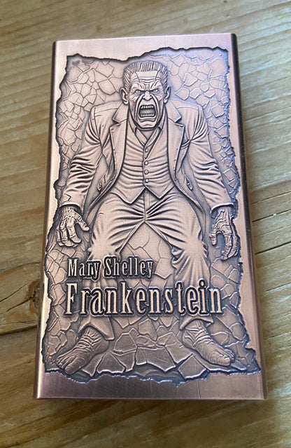 A rectangular metal box featuring an embossed illustration of Mary Shelley as Frankenstein, placed on a wooden surface. The box has a metallic rose-gold or copper-toned finish with gray engraved artwork and the text 'Mary Shelley Frankenstein' on the front.
