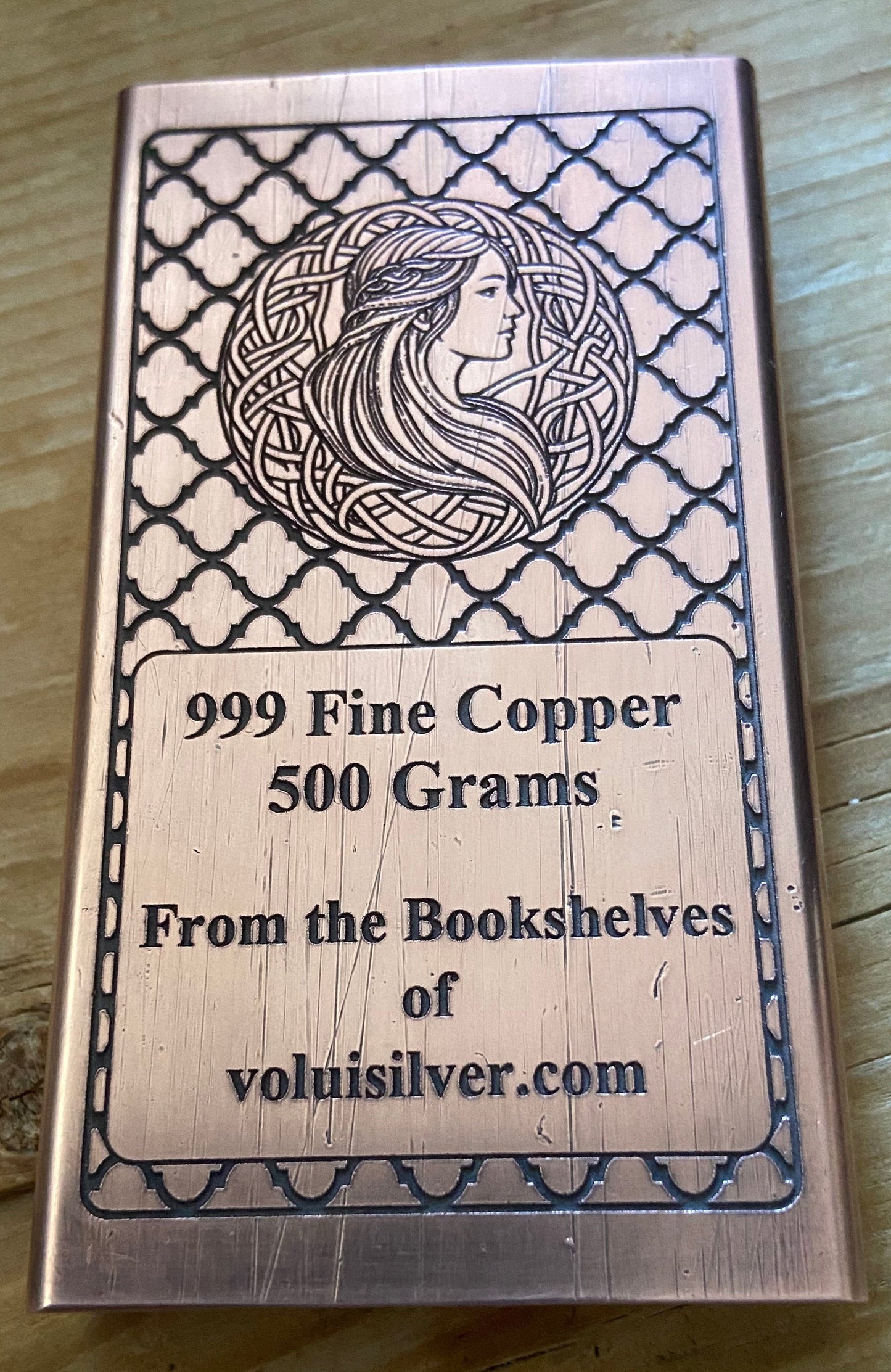 A rectangular copper ingot with engraved Celtic-style border patterns and a profile illustration of a woman, placed on a wooden surface. The text on the ingot reads '999 Fine Copper 500 Grams.