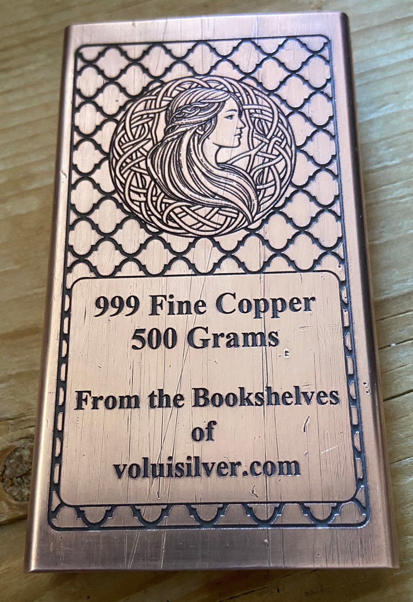 A rectangular copper ingot with engraved Celtic-style border patterns and a profile illustration of a woman, placed on a wooden surface. The text on the ingot reads '999 Fine Copper 500 Grams.