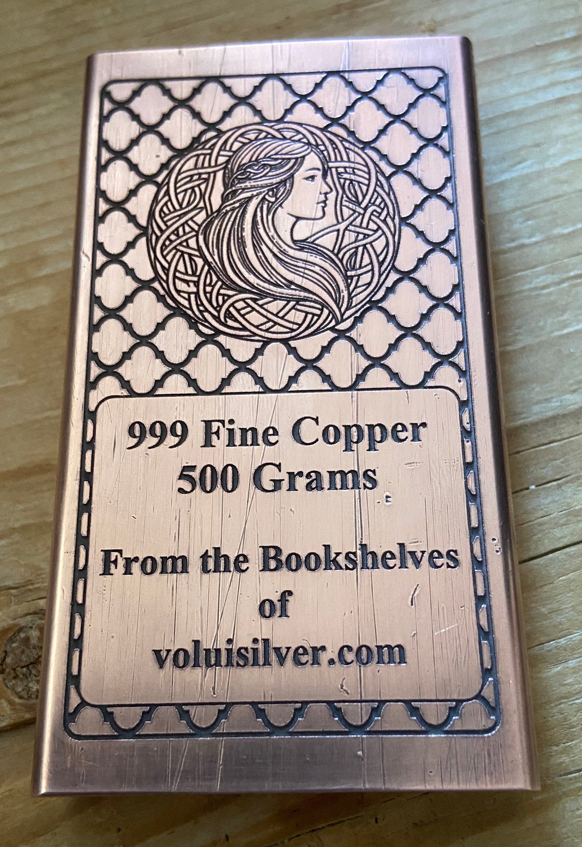 A rectangular copper ingot with engraved Celtic-style border patterns and a profile illustration of a woman, placed on a wooden surface. The text on the ingot reads '999 Fine Copper 500 Grams.