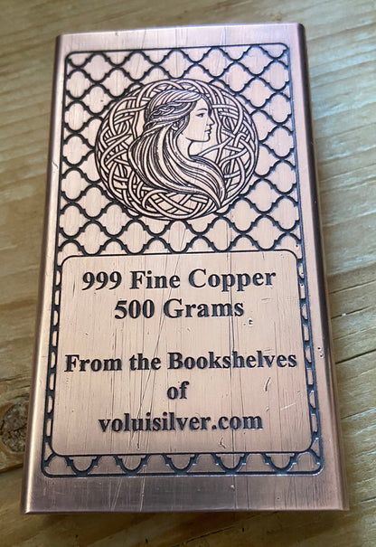 A rectangular copper ingot with engraved Celtic-style border patterns and a profile illustration of a woman, placed on a wooden surface. The text on the ingot reads '999 Fine Copper 500 Grams.