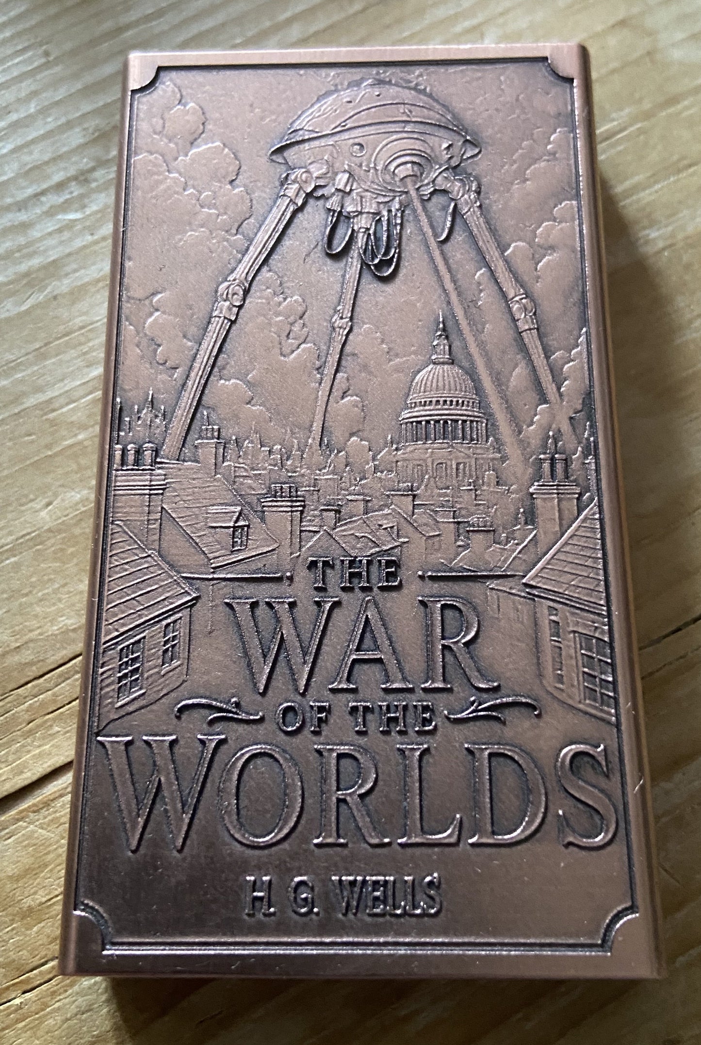 A rectangular bronze-colored book or box cover titled "The War of the Worlds" by H. G.