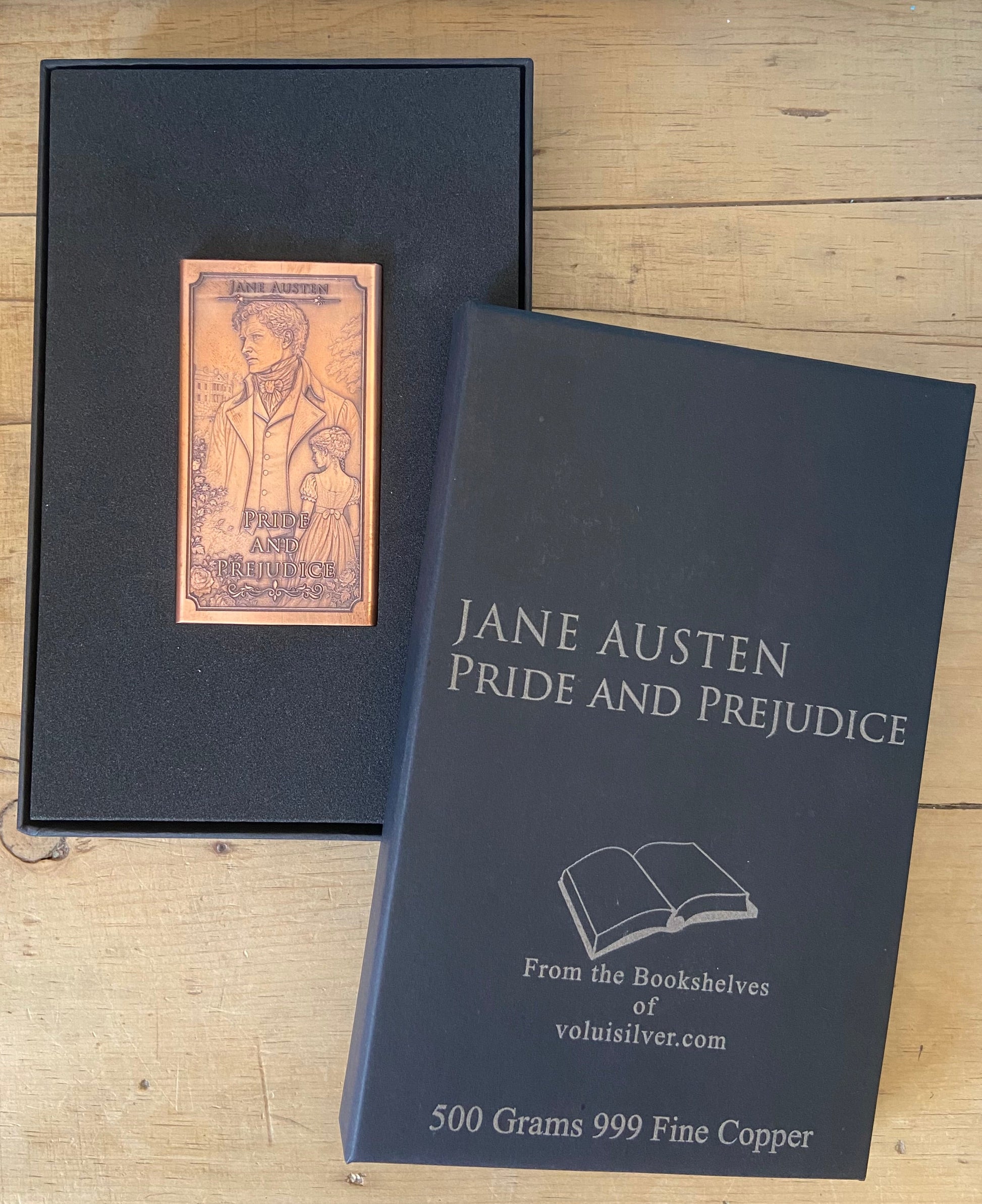 A Jane Austen Pride and Prejudice copper plate plaque displayed next to its black presentation box on a light wood background. The plaque is made of 500 grams of 999 fine copper and features an engraved portrait of Jane Austen.