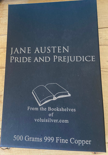 A rectangular black metal coin or medallion cover for Jane Austen's Pride and Prejudice, featuring white text and an illustration of an open book, placed on a light wood surface. The text reads 'From the Bookshelves of voluisilver.