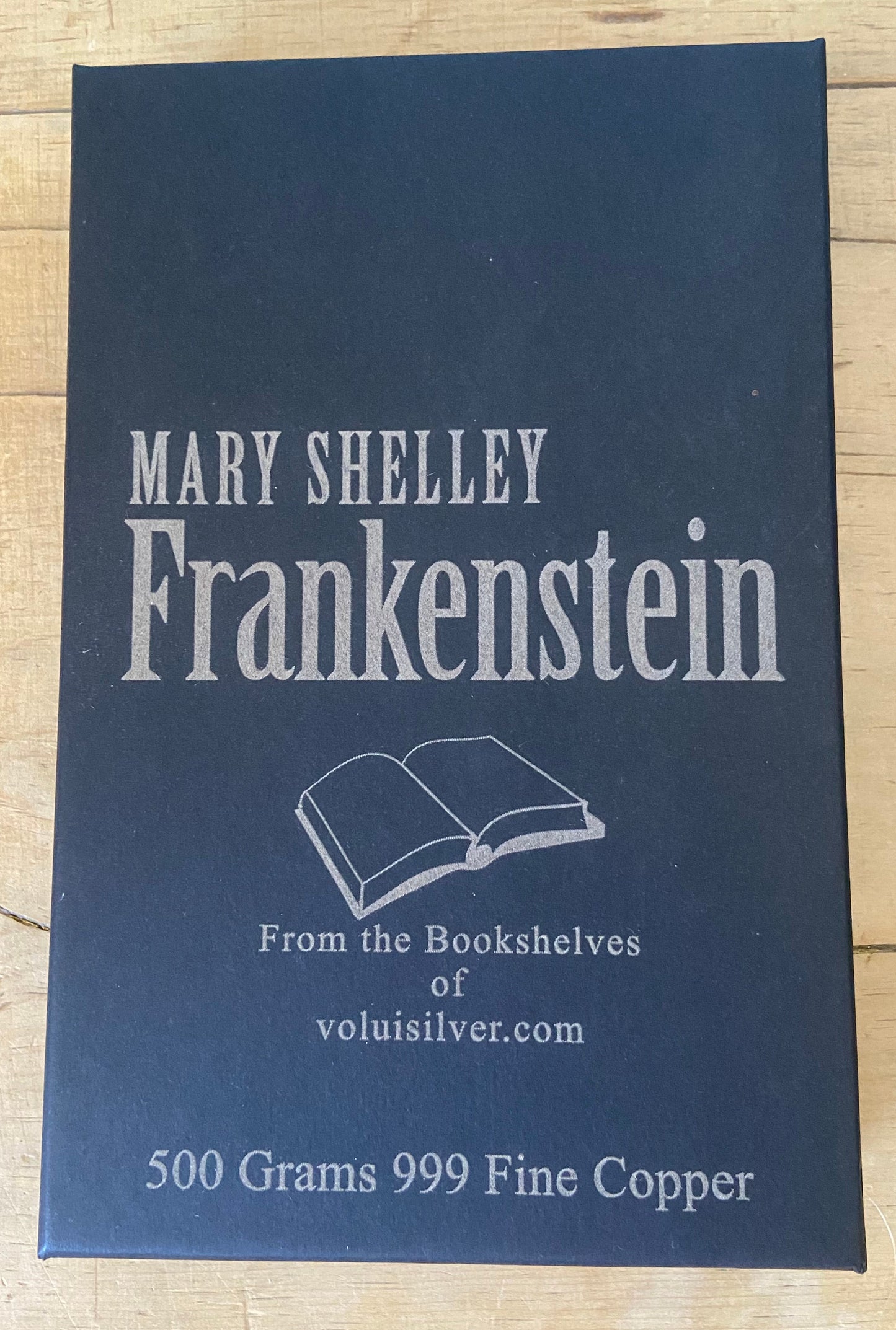 A rectangular dark blue metal tin labeled 'Mary Shelley Frankenstein' with white text and an open-book illustration, placed on a light wood surface. The tin also reads 'From the Bookshelves of voluisilver.