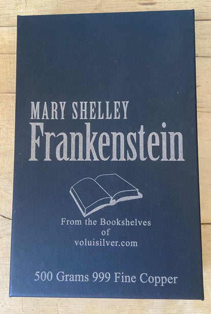 A rectangular dark blue metal tin labeled 'Mary Shelley Frankenstein' with white text and an open-book illustration, placed on a light wood surface. The tin also reads 'From the Bookshelves of voluisilver.