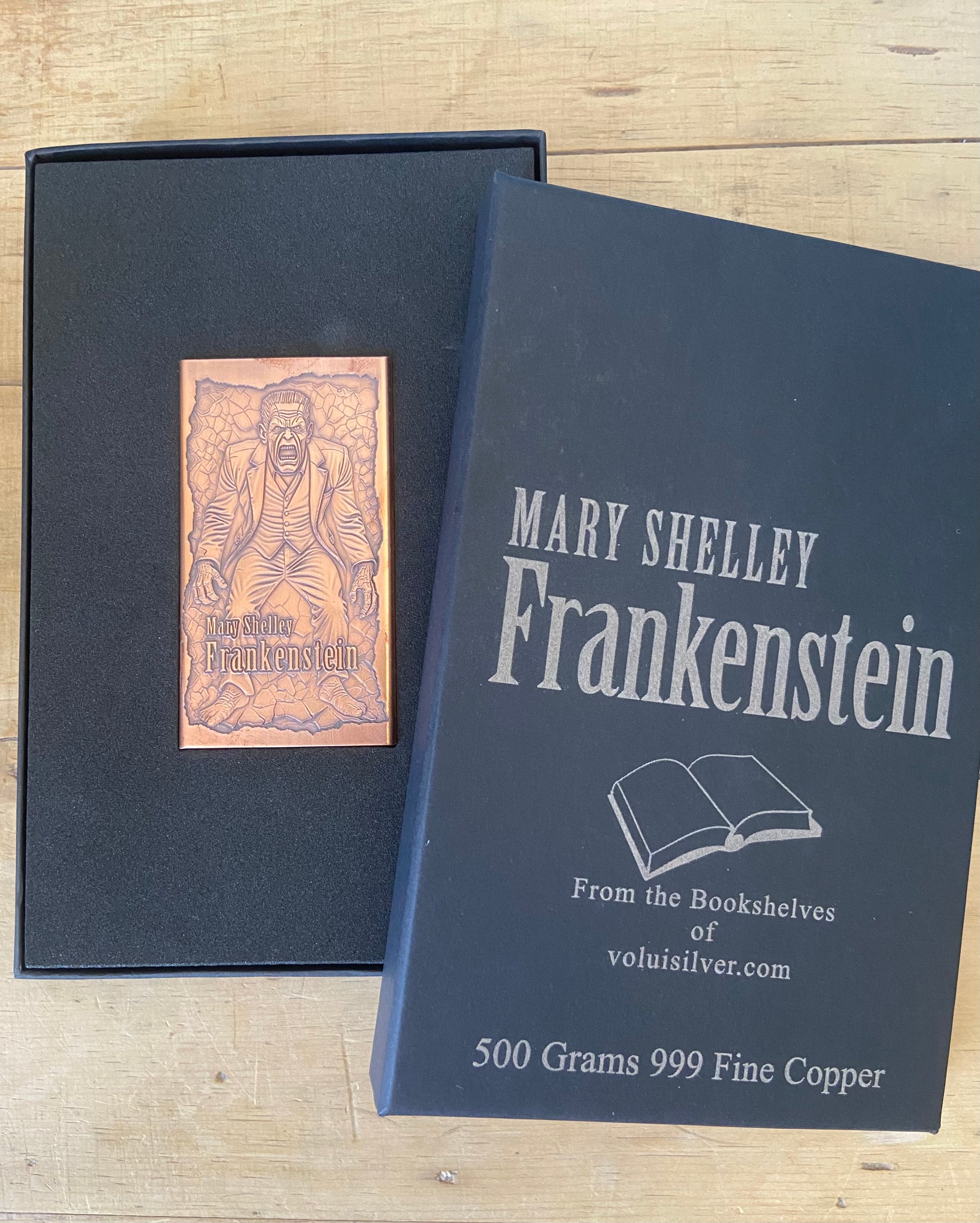 A copper plaque featuring an engraved illustration of Dr. Jonathan Crawford from Mary Shelley's Frankenstein, displayed next to its black presentation box on a light wood background.