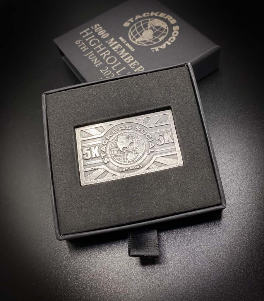 A 999 fine silver Stackers Social 5K displayed inside an open black presentation box, with the matching closed box and an engraved note behind it on a dark surface.