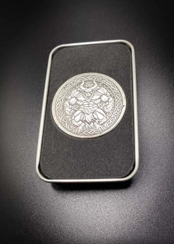 A silver-toned circular metal medallion featuring a winged demon or gargoyle figure, displayed inside a black rectangular case with a silver border on a dark background.