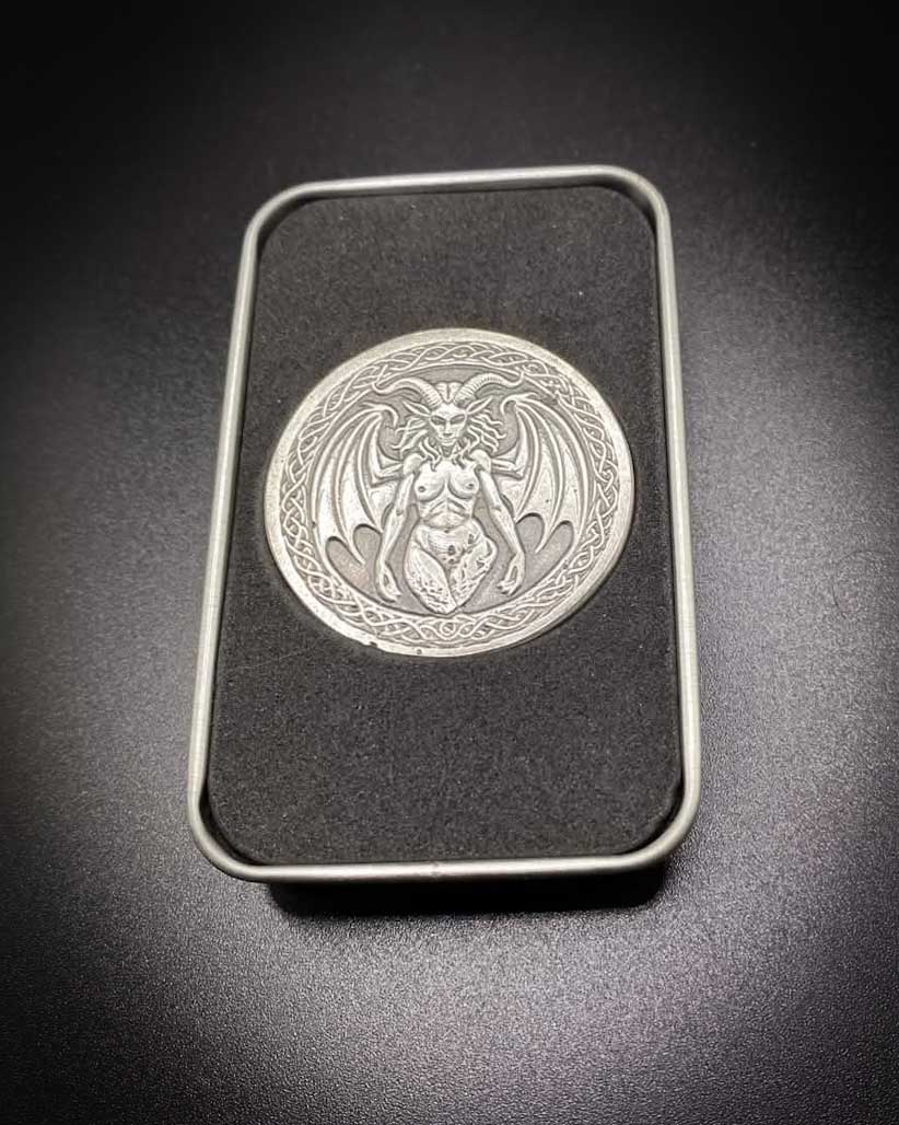 A silver-toned circular metal medallion featuring a winged demon or gargoyle figure, displayed inside a black rectangular case with a silver border on a dark background.